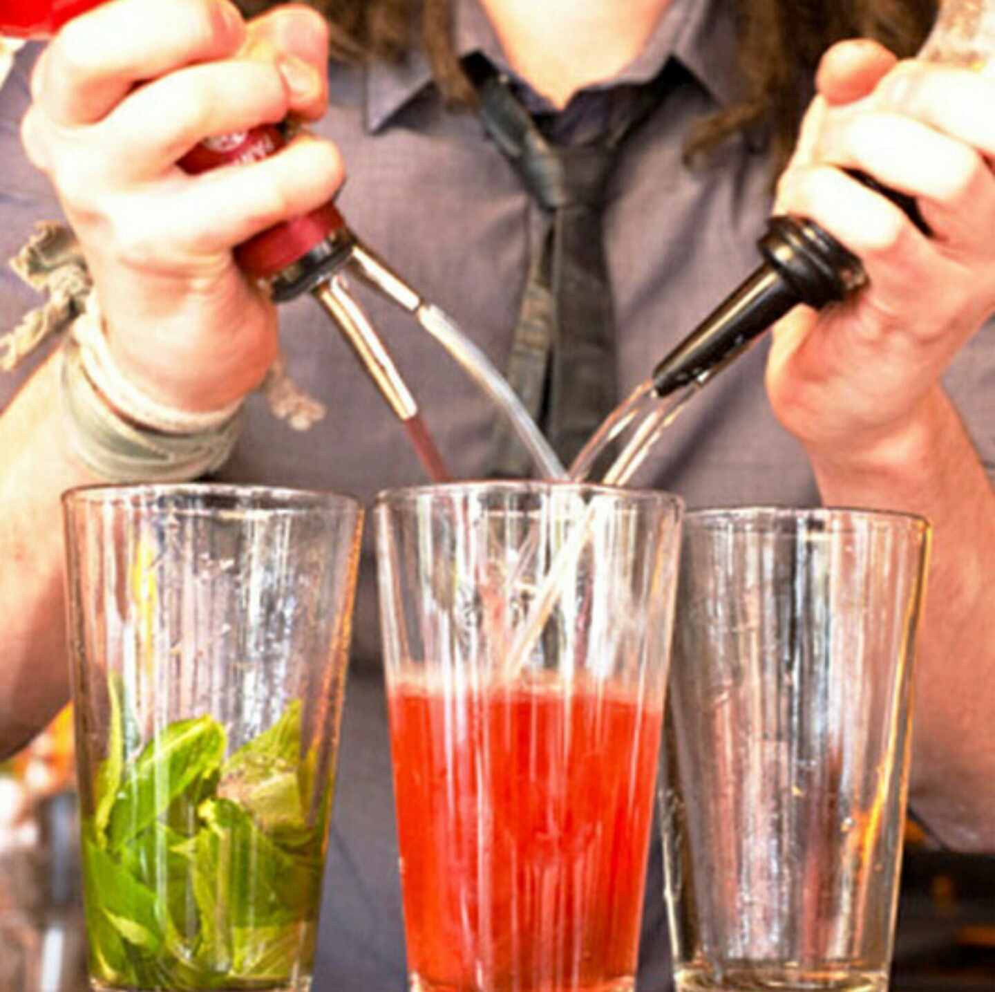Four bottles with speed pourers held with two hands pouring a mixed drink